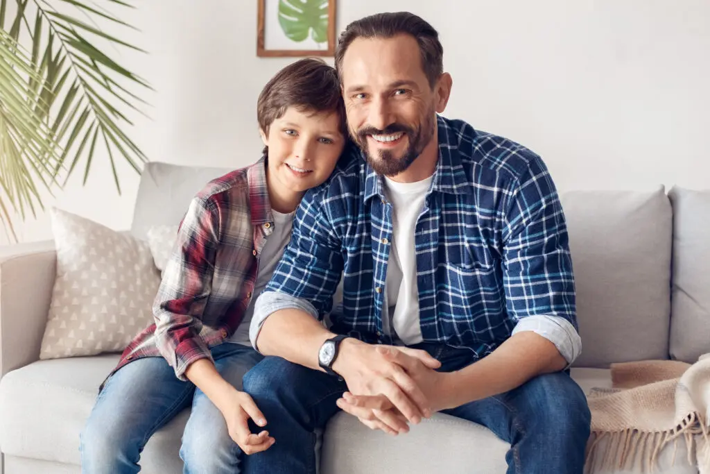 A father and son smile while sitting on a light gray sofa together