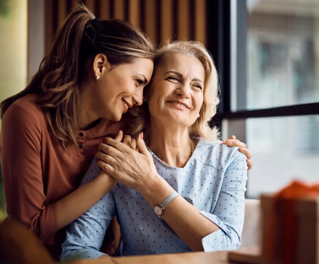 A woman lovingly embraces her mother, who sits at a table and glances out a window