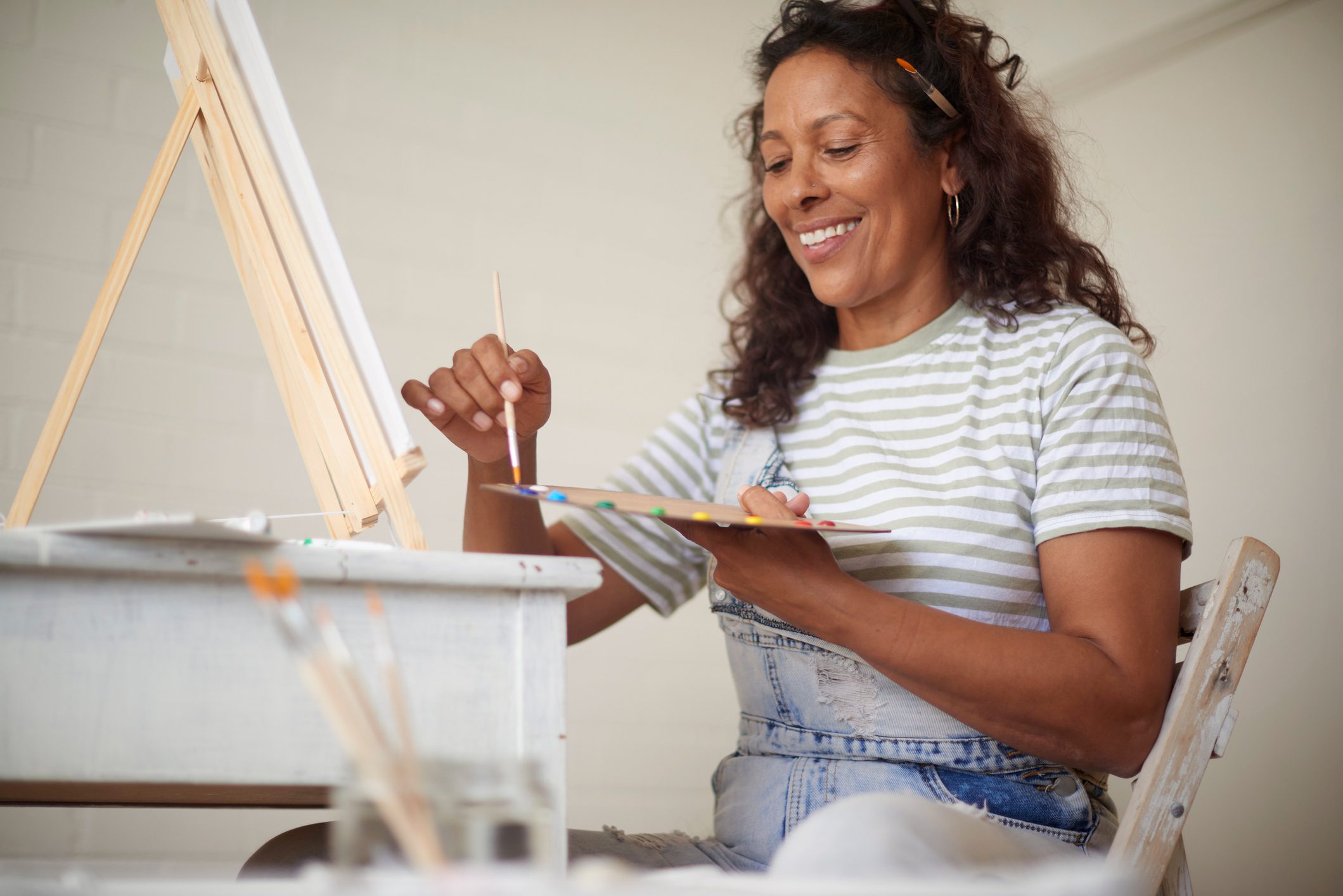 Woman smiles as she dips her brush into paint as she sits before a large canvas